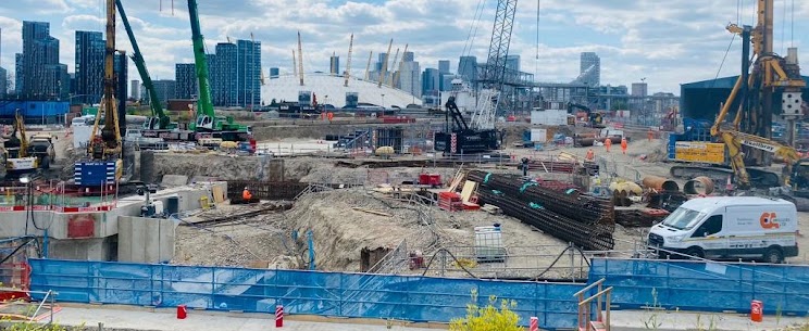 A bustling construction site in London featuring heavy machinery, cranes, and excavation work, with the iconic Millennium Dome (The O2 Arena) and Canary Wharf skyline visible in the background under a cloudy sky.